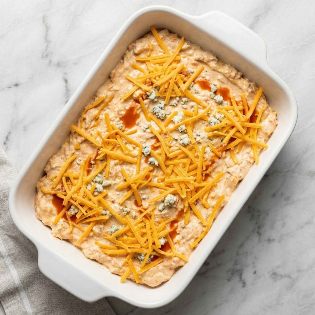 buffalo chicken dip mixture topped with shredded cheddar cheese in a baking dish before baking